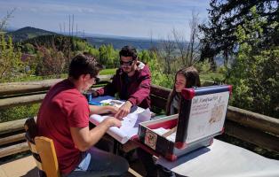 Drei Jugendliche sitzen draußen an einem Tisch in der Sonne und arbeiten mit Papier und einem Moderationskoffer.